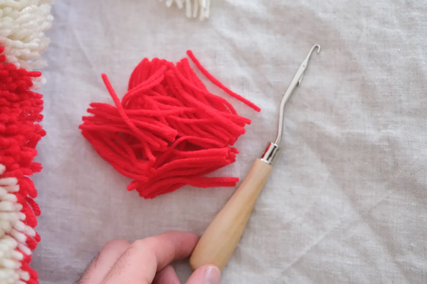 A hand holding a latch hook tool with red yarn, demonstrating the technique on a canvas