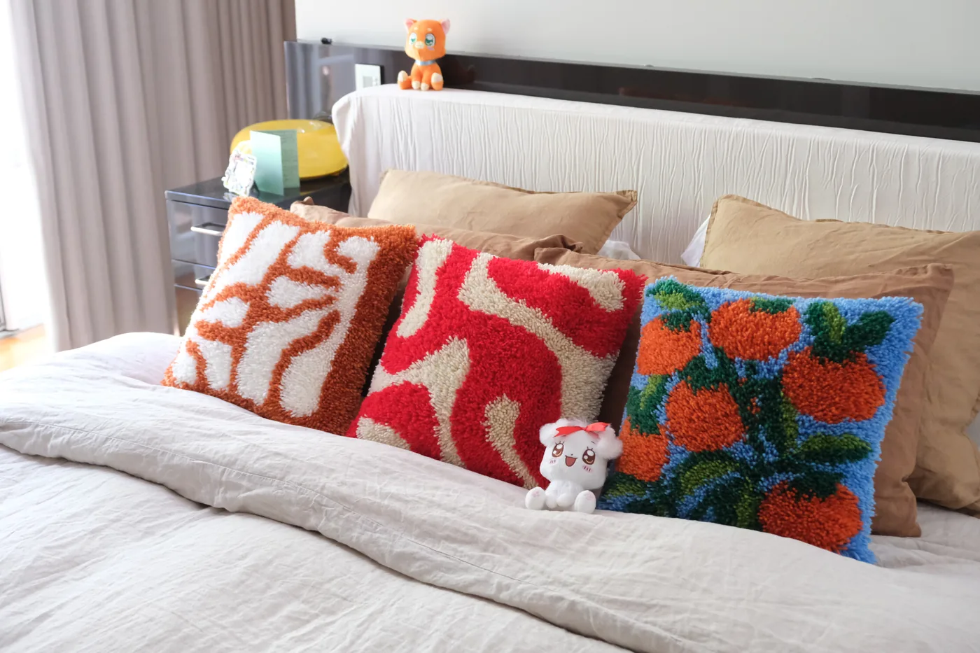 Three finished handmade latchhook cushions in different designs — terracotta, red and white stripe, and blue citrus — arranged on a white bed